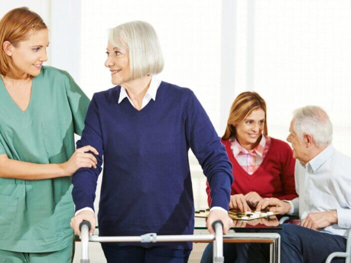 older woman with walker and nurse looking at each other, older man playing chess with women in the back