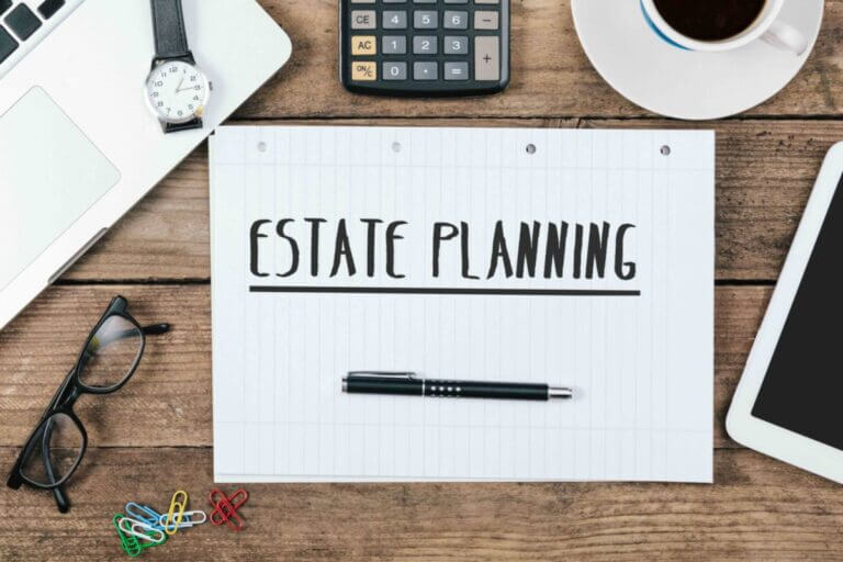 Estate Planning text on note pad, office desk with electronic devices, computer and paper, wood table from above