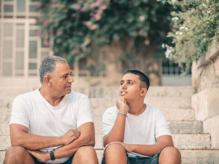 Teenager son and senior father sitting on stairs outdoors at home, talking.