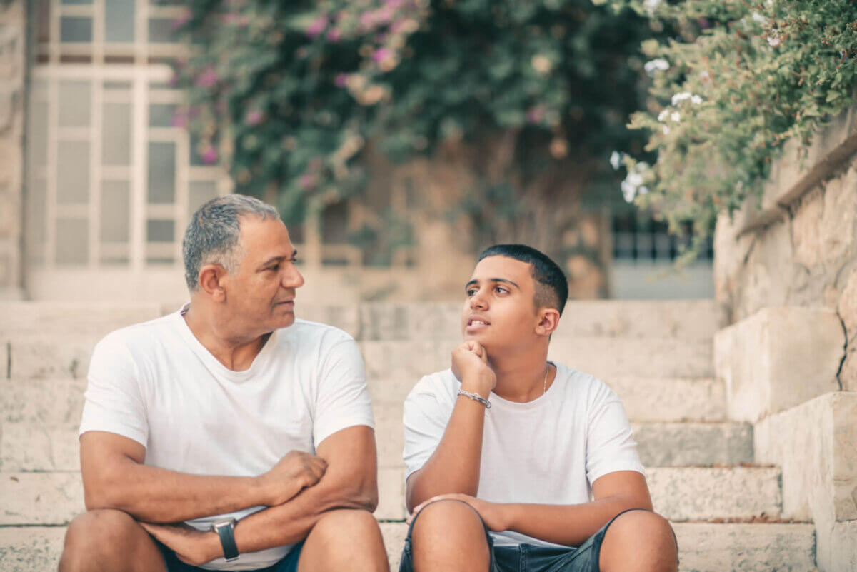 Teenager son and senior father sitting on stairs outdoors at home, talking.