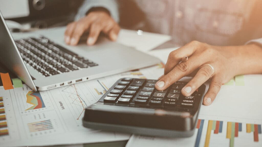 person on computer with calculator and spreadsheet next to them