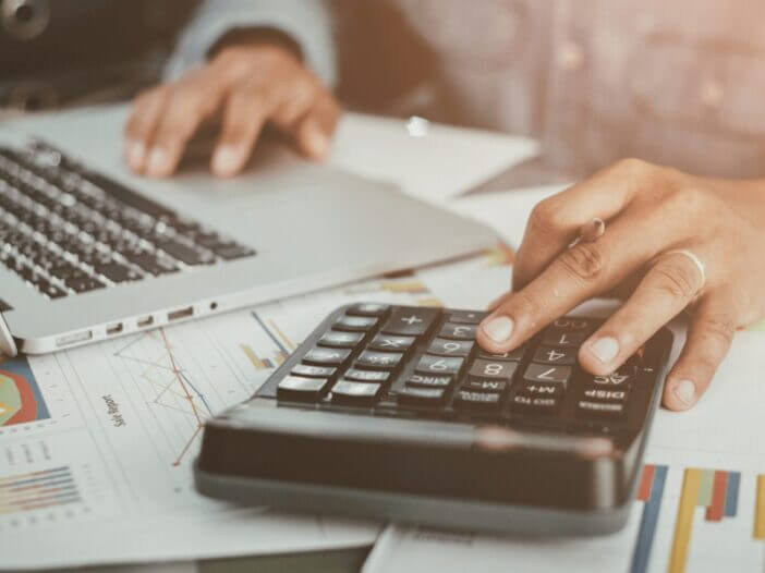 person on computer with calculator and spreadsheet next to them