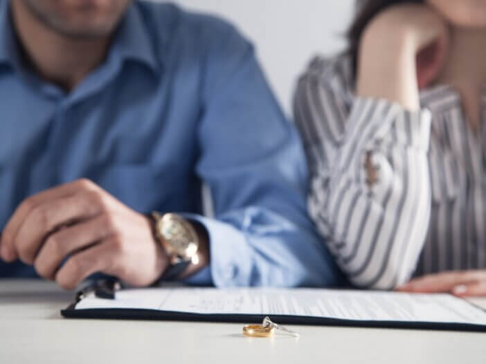 a picture of two wedding rings- one golden band and one diamond band and there is a couple in the background with divorce paperwork.