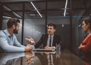 a lawyer sitting at a table while to the right of him there is a woman crossing her arms and to the left of him there is a man that looks upset.