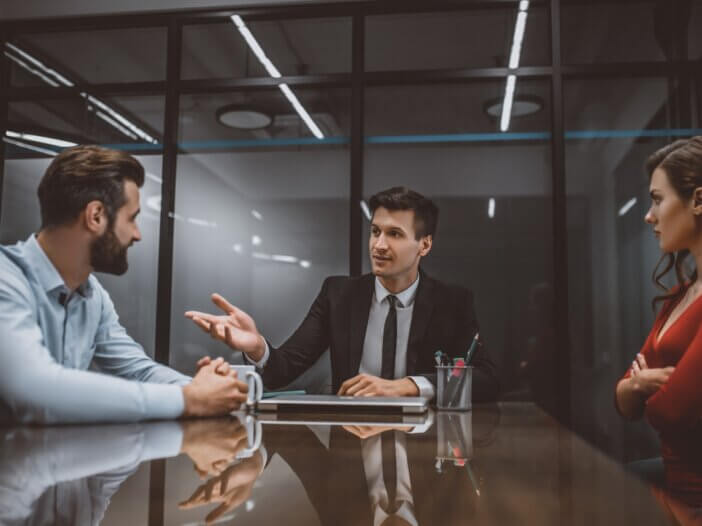 a lawyer sitting at a table while to the right of him there is a woman crossing her arms and to the left of him there is a man that looks upset.