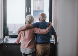 an older couple hugging in their kitchen. it is a photo of the elderly couple from behind and they are looking out their window.