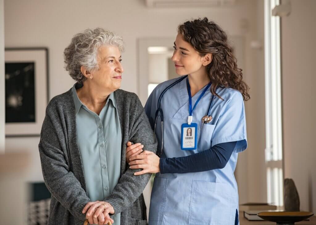 a caregiver and her patient walking down the hallway. the caregiver is on the right and the patient is on the left and they are looking at each other smiling.