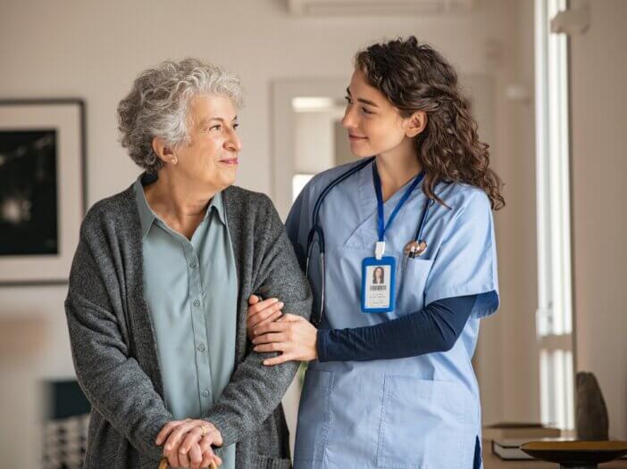 a caregiver and her patient walking down the hallway. the caregiver is on the right and the patient is on the left and they are looking at each other smiling.