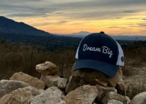 a picture of a snyder law hat- with the saying dream big up on rocks in the mountain and the sunrise in the back.