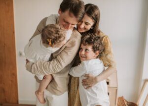 a mother and a father holding their two younger kids. the dad is holding the younger daughter and he is looking at here smiling and then the brother is hugging the mother who is smiling at him