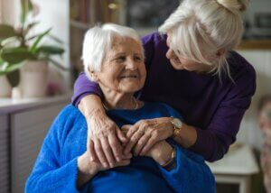 an older mother with her daughter. the mother is in a wheel chair while her daughter is behind hugging her shoulders. they are smiling