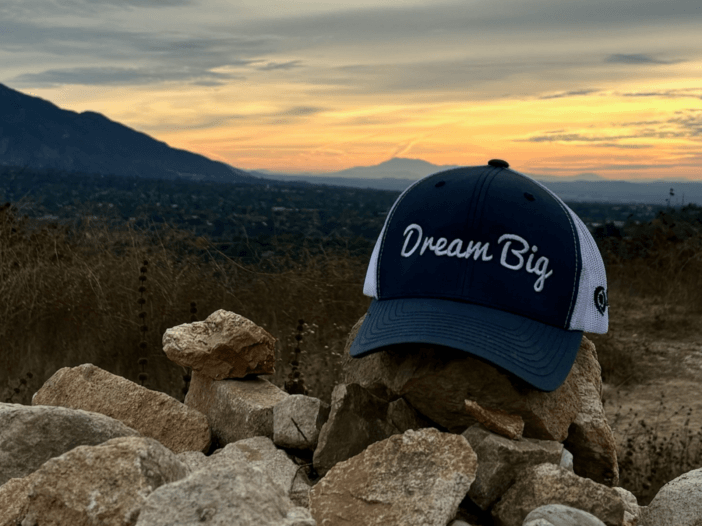 a picture of a snyder law hat- with the saying dream big up on rocks in the mountain and the sunrise in the back.