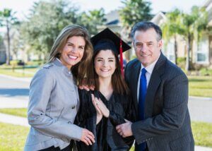 a young adult at their graduation with their parents. the graduate is in the middle, and then mom and dad are hugging and smiling with their daughter.