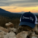 a picture of a snyder law hat- with the saying dream big up on rocks in the mountain and the sunrise in the back.