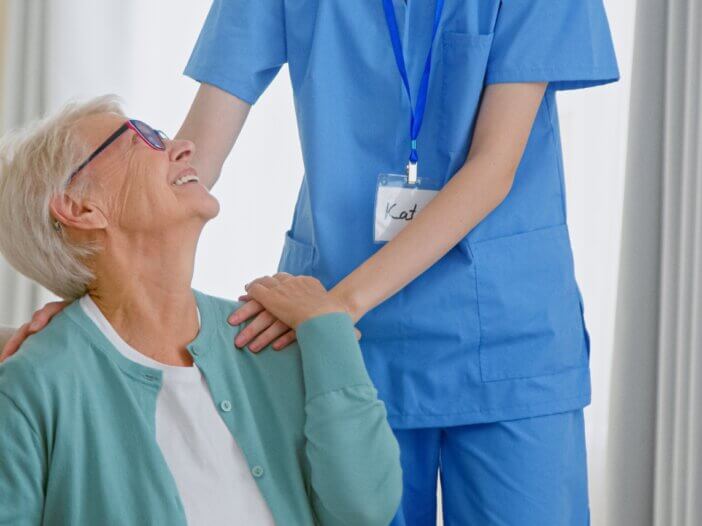 an older woman sitting in a chair and above her, you see a nurse putting her hands on her patients shoulders. the woman is looking up and smiling at the nurse.