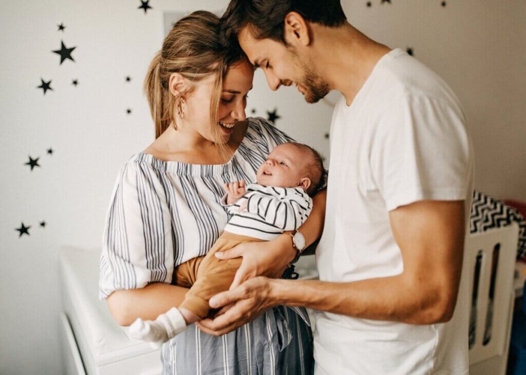 a mother and a father holding their newborn child. in the background is the babies crib and decorated bedroom.