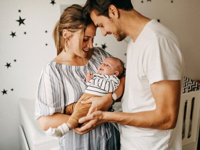 a mother and a father holding their newborn child. in the background is the babies crib and decorated bedroom.