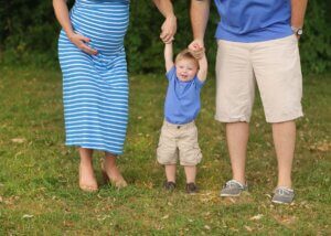 a little boy and his parents holding his hand on each side of them. you can see the parents torso and see that the mother is pregnant. the kid is in the middle smiling and swinging.