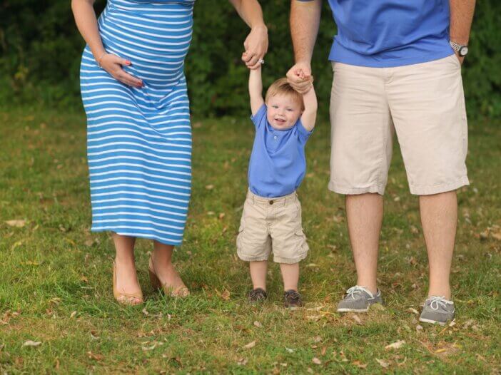 a little boy and his parents holding his hand on each side of them. you can see the parents torso and see that the mother is pregnant. the kid is in the middle smiling and swinging.