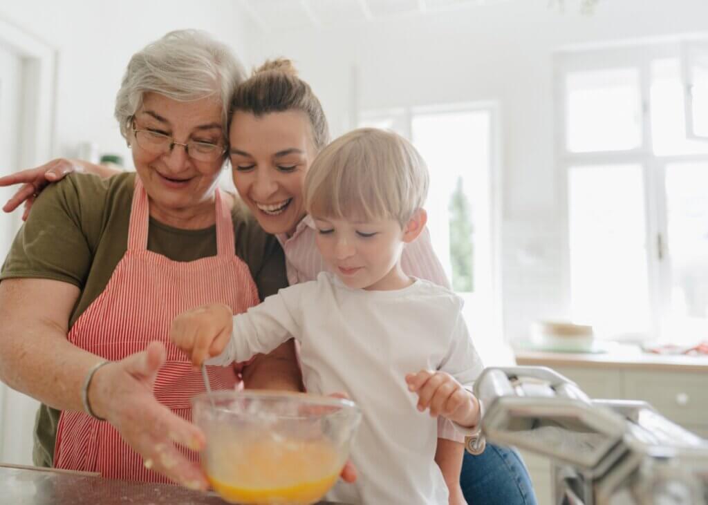 a grandmother and a mother baking with a little boy. they are all close together and smiling and mixing a bowl of ingredients.