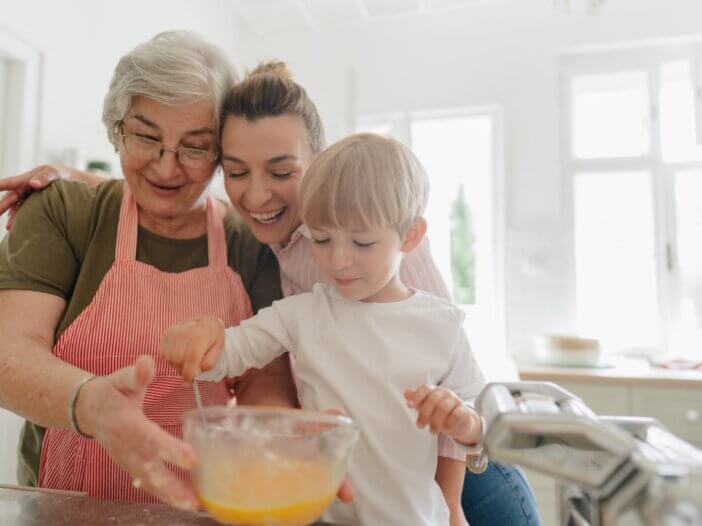 a grandmother and a mother baking with a little boy. they are all close together and smiling and mixing a bowl of ingredients.