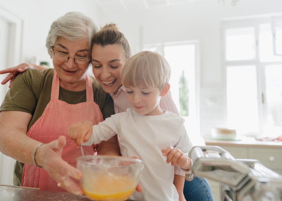a grandmother and a mother baking with a little boy. they are all close together and smiling and mixing a bowl of ingredients.