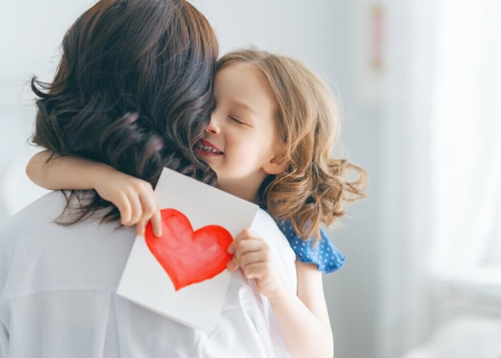 a mom and child hugging one another. the mothers back is turned and the daughter is hugging her and holding a heart.