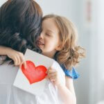 a mom and child hugging one another. the mothers back is turned and the daughter is hugging her and holding a heart.