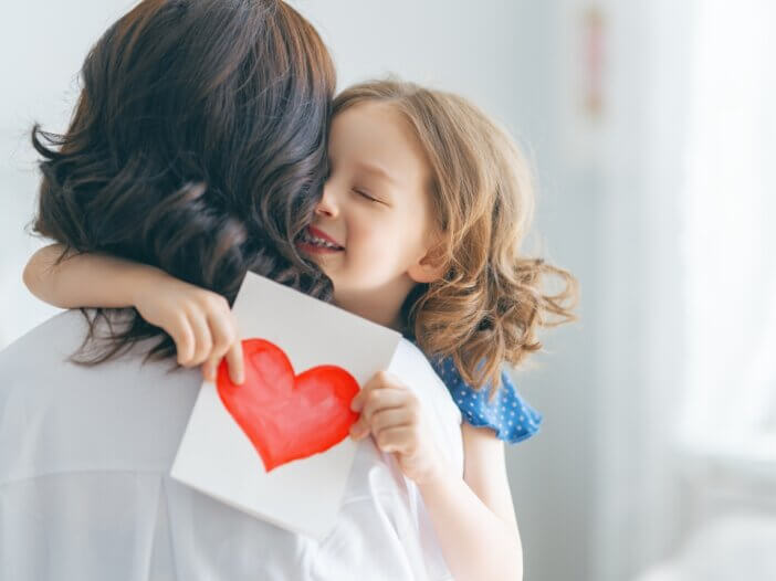 a mom and child hugging one another. the mothers back is turned and the daughter is hugging her and holding a heart.