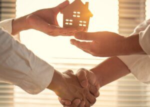 a person holding a small wooden estate shaking hands with someone and passing off that wooden home to them
