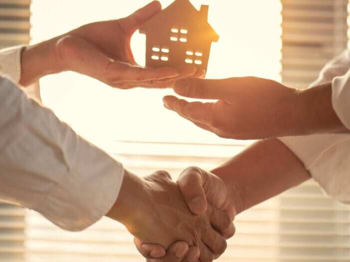 a person holding a small wooden estate shaking hands with someone and passing off that wooden home to them
