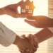 a person holding a small wooden estate shaking hands with someone and passing off that wooden home to them