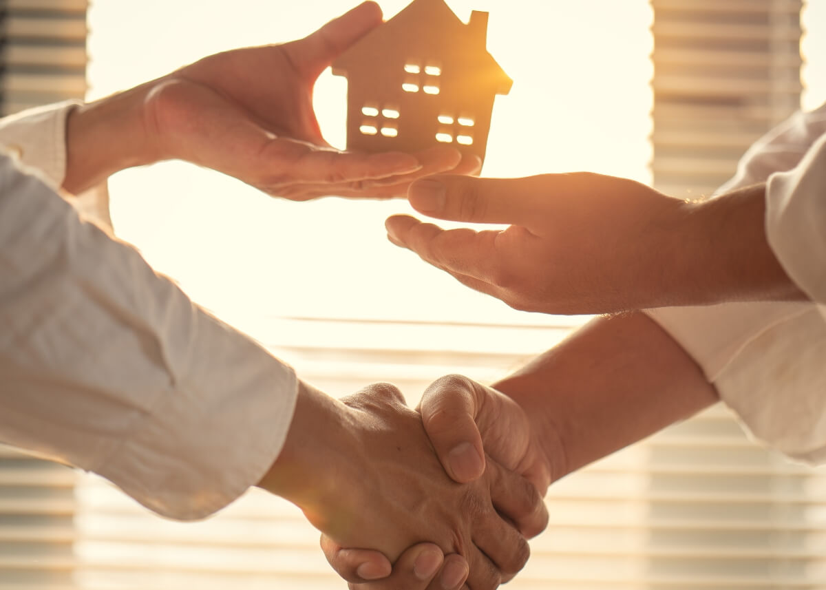 a person holding a small wooden estate shaking hands with someone and passing off that wooden home to them