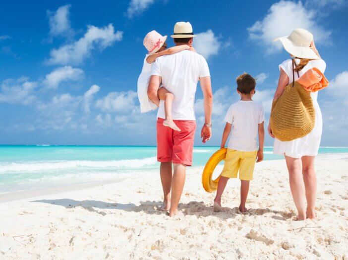 a family of four walking on the beach on vacation. it is a shot from behind with the mom on the right holding their sons hand and then the dad on the left holding the younger daughter