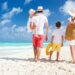 a family of four walking on the beach on vacation. it is a shot from behind with the mom on the right holding their sons hand and then the dad on the left holding the younger daughter