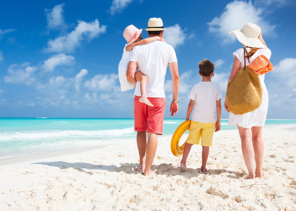 a family of four walking on the beach on vacation. it is a shot from behind with the mom on the right holding their sons hand and then the dad on the left holding the younger daughter