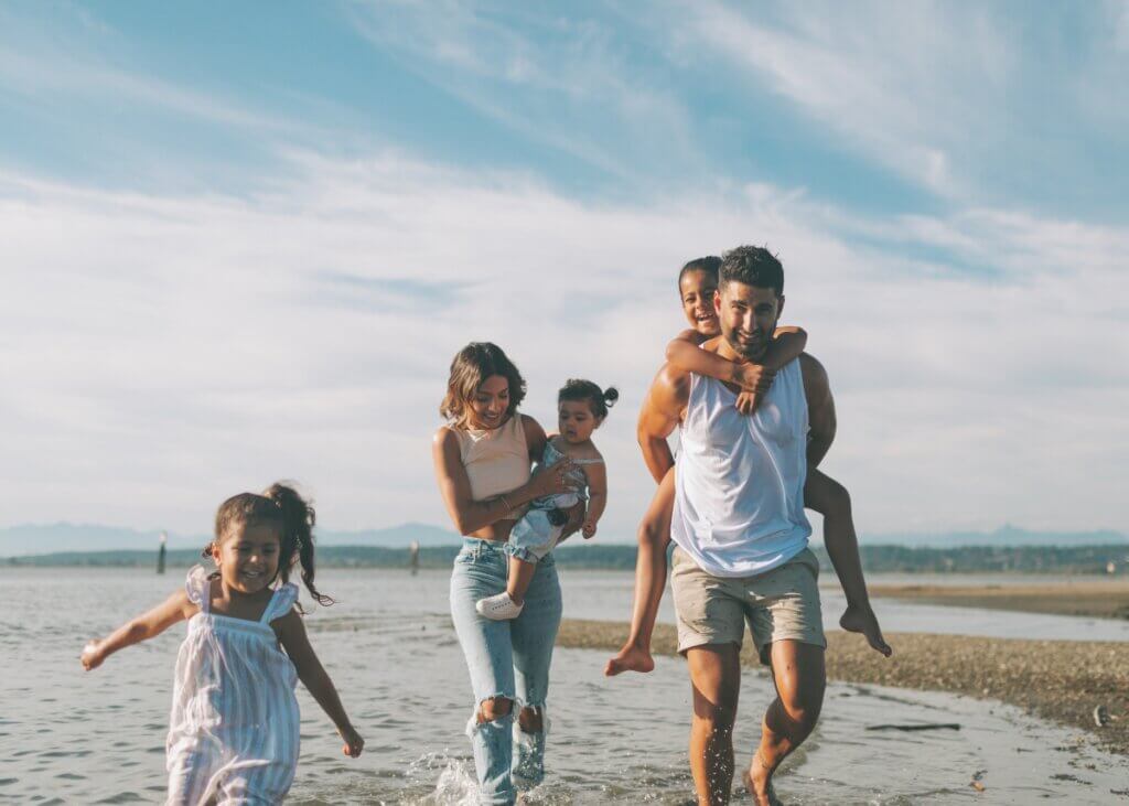 a family at the beach. there are three kids and the two parents with a mom holding one kid and a kid on the dads back.