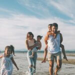 a family at the beach. there are three kids and the two parents with a mom holding one kid and a kid on the dads back.