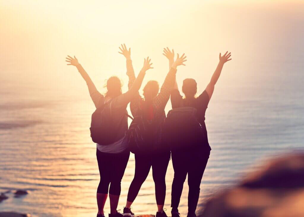 a picture of three people from behind on a mountain at sunset raising their hands up