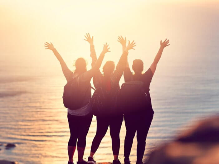a picture of three people from behind on a mountain at sunset raising their hands up