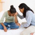 a mom sitting on her couch and consoling her daughter that is crying