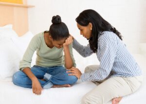 a mom sitting on her couch and consoling her daughter that is crying